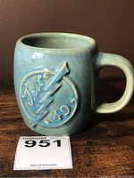 Vintage four inch majolica mug with raised circular medallion featuring 'Test 49' and lightning bolt design on front. Light blue glaze with rounded handle and body.