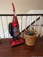 Red Bissell Cleanview II bagless upright vacuum with cord wrapped and clear dirt container, standing next to a wicker basket filled with metal hangers, placed on dark hardwood floor against white stair railing.