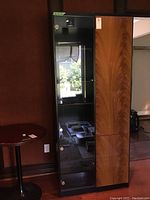 Front and side view of tall wood and glass cabinet with glass door on the left and wood door on the right, interior shelving visible through glass door.