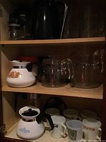 Shelf displaying white ceramic coffee carafes with floral patterns and multiple vintage mugs below