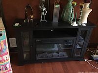 Front view of black electric fireplace TV stand showing central fireplace insert with faux logs and two glass-door storage cabinets on each side, decorated with various ornamental items on top.
