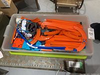 Top view of plastic container filled with orange Hot Wheels track pieces, blue and white connectors, launcher parts, and multiple Hot Wheels cars.