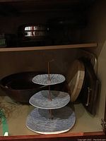 Photo of three-tier serving platter with three textured ceramic plates and metal handle inside cabinet with wooden bowls and cutting boards behind