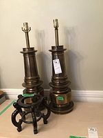 Photo of two tall brass-colored metal lamp stands side by side with bulb sockets at the top, standing on a floor against a wall.