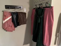 Six skirts hung on hangers against wall: pink fabric skirts, black leather skirts, brown leather skirt, dark green suede skirt with belt.