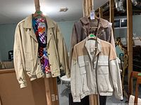 Three leather jackets hanging on hangers on a wooden rack in a basement. Left is Bilermain cream jacket with colorful lining, middle beige jacket with tan panels, right brown jacket with worn collar.