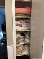 Linen cupboard with various folded linens and blankets stacked on shelves, including pink and green wool blankets on the top shelf and assorted white and floral linens below.
