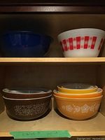 Shelved display showing four Pyrex bowls: dark blue solid bowl, white bowl with red checkered pattern, brown bowl with white floral decoration, and three orange bowls with white floral designs stacked.