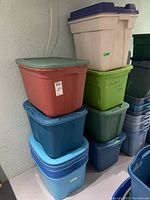 Seven stacked plastic storage containers with various colored lids in a basement setting.