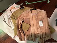 Three ladies jackets displayed on hangers laid on the floor: light pink perforated suede jacket, beige suede jacket with smocked detailing, and white leather jacket.