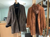 Photo of two suede coats hanging side by side; dark brown coat on left with black fur lining, tan coat on right with a reddish-brown fur collar