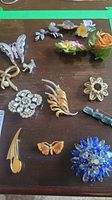 Full view of various vintage brooches on dark wood surface including floral and butterfly designs with rhinestones and enamel.