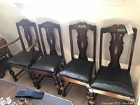Photo shows a set of four wooden dining chairs in a row, with dark wood frames and black padded seats; one has armrests, others do not; placed indoors against a white wall.