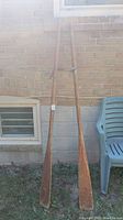 Full view of two wooden oars propped against a brick wall and a plastic chair, showing overall shape and condition.