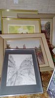 Five framed prints stacked and leaning against a wall, showing one black and white architectural print and four colored landscape/flower prints.