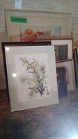 Multiple framed artworks leaning against a wall, including floral prints and watercolors.