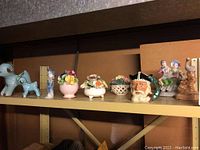 Full shelf view of the lot showing all figurines including Royal Doulton Viking toby jug, floral baskets, Japanese figures, lambs, and ducks.