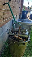 Green plastic garbage pail filled with assorted scrap metal, old motor, tire iron, and garden tools against brick wall outside