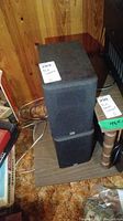 Photo of two stacked black PSB Alpha Mini speakers with front grilles, placed on a board against a wooden wall.