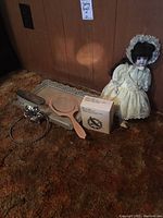 Sterling brush, two mirrors, boxed binoculars, silver-tone binocular neck chain, and a vintage doll dressed in yellow/cream lace dress with bonnet on brown carpet floor.