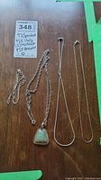 Four pieces of 925 sterling silver jewelry arranged on wooden surface. Two simple chains, one pendant chain with green stone, and one three-strand bracelet.