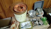 Full lot of vintage baking supplies including tins, cake pans, mixing bowls, sifter, cookie cutters and measuring cup on wood-paneled table.