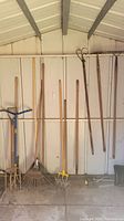 Photo of multiple garden tools arranged upright on a shed wall showing shovels, pitchforks, rakes, aerator, and sprinkler head.