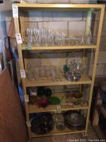 Metal utility shelf with four shelves shown against a concrete block wall, holding various glassware items (not included) in basement.
