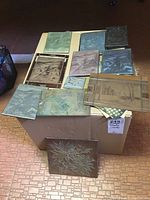 Top-down view of 10 metal printing plates laid on a box, displaying various detailed etched designs with some visible signatures and Christmas themes.