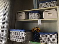Photo showing four white wicker baskets with blue fabric linings on shelves and two beige cardboard storage boxes with lids.