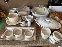 Multiple white ceramic and CorningWare kitchen items including casseroles, ramekins, condiment bowls, muddler, and serving bowl basket