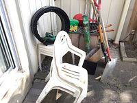 White resin stackable chairs, black garden edging roll, various long-handled garden tools including shovels and a rake, and other garden accessories placed near a wall.
