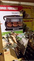 Photo showing George Foreman grill in its original box with grilling surface visible, two heating discs packaging behind, and multiple BBQ tools including spatulas and tongs in a metal holder.