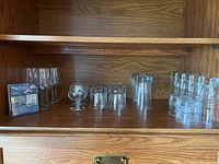 Shelf with all glassware and package of wine charms displayed, including wine flutes, brandy snifter, scotch glasses, tumblers.