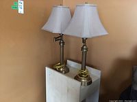 Two matching brass-finish table lamps with white shades placed on a small white rolling table against a brown wall.