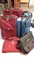 Photo showing two red soft-sided luggage suitcases and one blue hard-shell suitcase with wheels, along with a smaller red travel bag and a black Timken laptop bag in front.