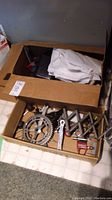 Cardboard box containing bicycle chainrings, cranks, pedals, and some painting supplies such as gloves.