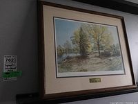 Framed print displayed, showing its size and overall look, a landscape with trees and river.