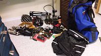 Wide view of assorted sports equipment on beige carpet including helmets, bags, shoes, hand pump, tennis racket, gloves, mask, and miscellaneous bicycle parts scattered around.