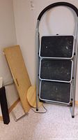 Three-step metal step stool leaning against wall with round wooden cutting board and rectangular wooden dollie beside it.