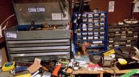 Metal tool box with drawers open showing assorted tools and workspace with other items on table surface