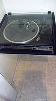 Technics turntable closed with transparent dust cover, showing platter and tonearm inside, positioned on tiled floor.