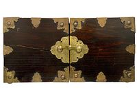 Top view of the closed jewelry box showing dark wood with brass corner and edge brackets and a central brass locking latch.