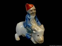 Porcelain figurine of leprechaun in red hat and blue clothes sitting on white pig, side angle showing details.