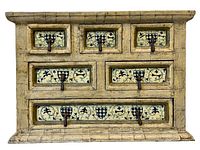 Front view of vintage painted jewelry box showing six small drawers and one long drawer. Details of painted floral and heraldic-like blue and black pattern on cream background, and metal pull rings visible.