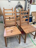 Four natural wood ladder back dining chairs arranged in a warehouse space, showing front and top views. Two chairs show paint splatter and wear marks on seats.