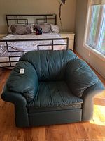Front angled view of green leather arm chair showing cushioned seat, armrests, and part of a bed in the background.