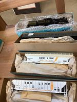 Photo showing black steam locomotive engine with tender in bubble wrap box, black passenger coach wrapped in paper, and one Soo Line white covered hopper car in box.