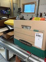 Boxed items stacked in a workshop environment showing a labeled banker's box and other boxes with model train components.