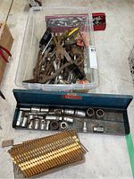 Plastic bin filled with assorted hand tools, wrenches, sockets, pliers, and nail gun nails, with a blue metal socket case in front and a coil of nail gun nails placed beside it on the floor.